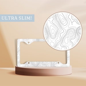 May include: White license plate frame with a topographic map design. The frame is ultra slim and has a modern look.