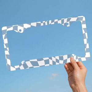 May include: A rectangular license plate frame featuring a repeating warped checkerboard pattern in shades of light blue and white. The frame has four circular mounting holes positioned at the corners to allow for attachment to a vehicle.