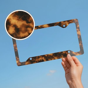 May include: A license plate frame with a tortoiseshell pattern. The frame is held against a blue sky. The frame has a rectangular shape with rounded corners and three holes for mounting. A circular inset shows a close-up of the tortoiseshell pattern.