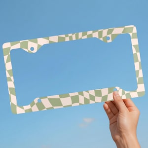 May include: A rectangular license plate frame featuring a wavy, distorted checkerboard pattern in alternating shades of sage green and cream. The frame includes four circular mounting holes positioned at the corners for attachment to a vehicle.