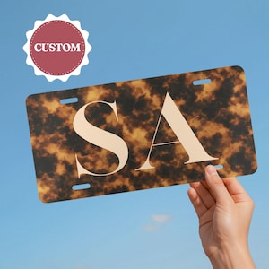 May include: A rectangular license plate with a tortoiseshell pattern and the letters "SA" in a cream color. The word "CUSTOM" is in a red and white circular badge in the upper left corner. The background is a clear blue sky.