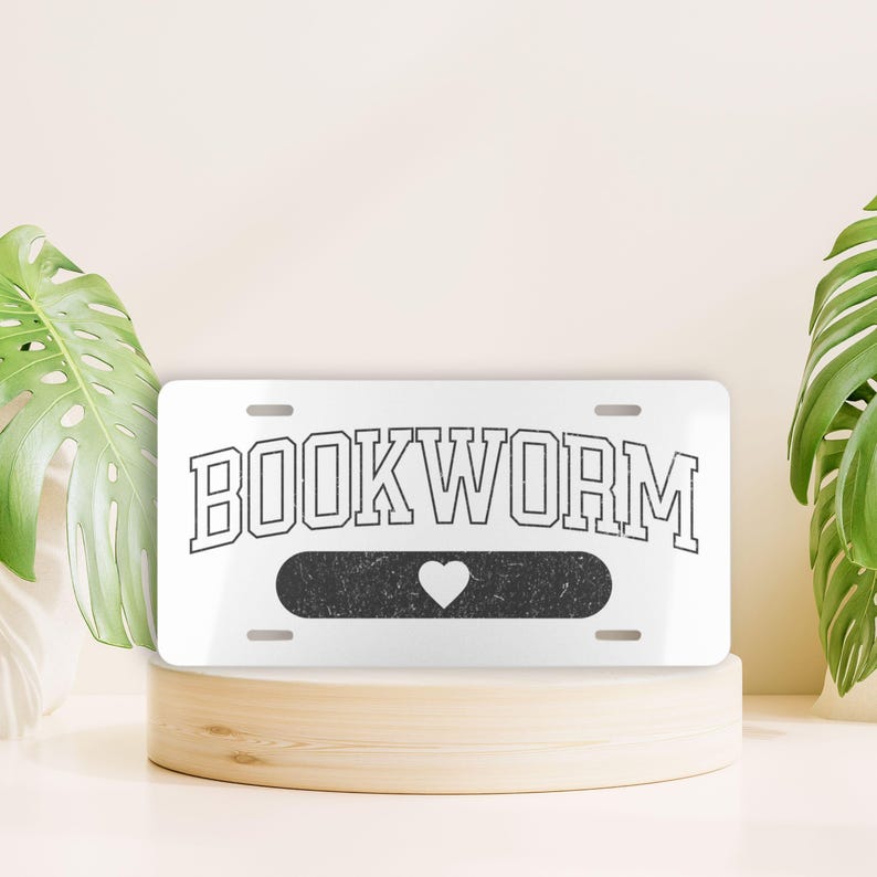 May include: White metal license plate with black text that reads "BOOKWORM" with a black rectangle underneath with a white heart in the centre.