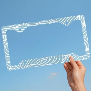 May include: A rectangular license plate frame featuring a light blue background with a repeating white line pattern of swirling waves. The frame has four circular mounting holes positioned at the top and bottom corners for vehicle attachment.