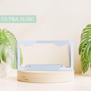 May include: A light blue license plate frame with rounded corners and two holes on each side. The frame is on a wooden platform and the text "ULTRA SLIM!" is visible in the top left corner.