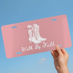 May include: A pink license plate featuring a white illustration of cowboy boots and the text "Walk By Faith" in a cursive font. The plate has two slots at the top and bottom. The background is a clear blue sky.