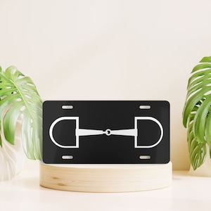 May include: Black and white metal license plate featuring a white silhouette of a horse bit. The license plate is on a wooden stand.