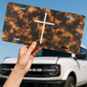 May include: License plate with a tortoiseshell pattern and a white cross. The plate has four mounting holes and is held up against a blue sky and a white vehicle.