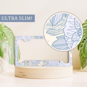 May include: A white license plate frame with a blue floral pattern. The frame is labeled "ULTRA SLIM!" and is shown on a wooden surface.