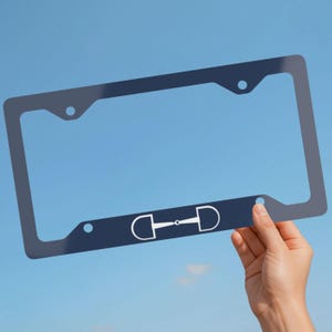 May include: A dark navy blue rectangular license plate frame featuring a white horse bit graphic centered on the bottom rail. The frame has four circular mounting holes, two at the top and two at the bottom, designed for standard vehicle license plates.