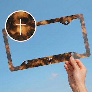May include: A license plate frame with a tortoiseshell pattern and white cross accents. The frame is held against a blue sky. A circular detail shows a close-up of the pattern and cross.
