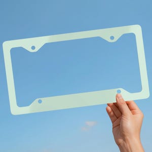 May include: A pale mint green rectangular license plate frame with rounded corners and four circular mounting holes, designed to hold a standard vehicle license plate. The frame features a smooth, solid-colored finish and a simple, open-center design.