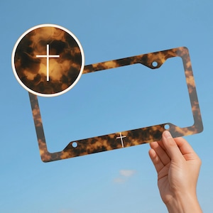 May include: A license plate frame with a tortoiseshell pattern and a white cross design. The frame is held against a blue sky. The frame has a circular inset with a cross and a cross at the bottom.