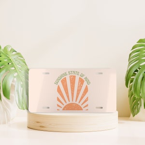 May include: A white metal license plate with a peach-colored sun graphic and the text "Sunshine State of Mind" in green.