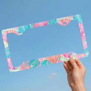 May include: A rectangular license plate frame featuring a repeating pattern of pink, coral, and turquoise seashells and coral reef elements against a white background. The frame has four circular mounting holes at the corners for attachment.