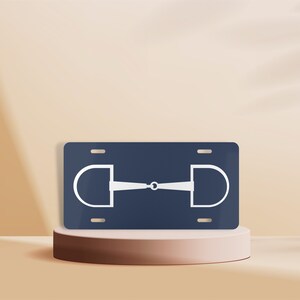 May include: A blue license plate with a white silhouette of a horse bit. The bit is a simple design with two rings and a center bar.