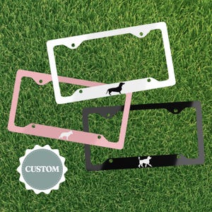 May include: Three rectangular number plate frames in white, pink, and black. The white frame features a black dachshund silhouette, the pink frame displays a white dog silhouette, and the black frame shows a white dog silhouette. A badge reads CUSTOM.