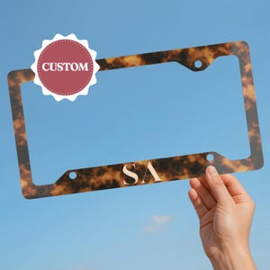 May include: A license plate frame with a tortoiseshell pattern. The frame has the initials "SA" in a gold color. A red and white circular sticker with the word "CUSTOM" is in the upper left corner. The frame is held against a blue sky.