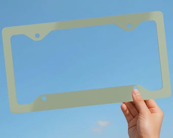 May include: A rectangular, sage green license plate frame with four circular mounting holes positioned at the corners. The frame features a smooth, solid-colored finish and a standard design intended to surround a vehicle license plate.