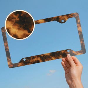 May include: A license plate frame with a tortoiseshell pattern. The frame is held against a blue sky background. The frame has a rectangular shape with rounded corners and three holes for mounting.