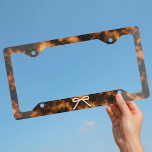May include: A rectangular license plate frame featuring a brown and black tortoiseshell pattern. The frame has four circular mounting holes and a small, centered, light-colored line drawing of a bow on the bottom horizontal edge.