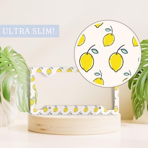 May include: White license plate frame with a lemon pattern. The frame is ultra slim and has a white background with yellow lemons and green leaves. "ULTRA SLIM!" is written in blue text on the top left corner of the image.