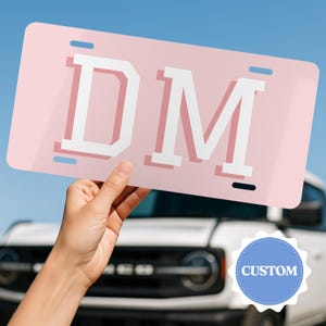 May include: A rectangular light pink license plate featuring the white capital letters DM in a bold varsity font with a subtle pink drop shadow. The plate includes four mounting slots at the top and bottom corners for attachment to a vehicle.
