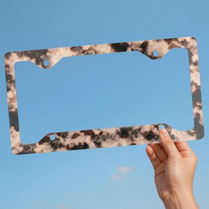 May include: A rectangular license plate frame featuring a mottled, tortoiseshell-inspired pattern in shades of dark brown, tan, and cream. The frame has four circular mounting holes positioned at the corners for secure attachment to a vehicle.