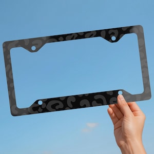 May include: A rectangular number plate frame featuring a dark grey and black leopard print pattern. The frame has four circular mounting holes positioned at the corners to allow for secure attachment to a vehicle's number plate area.