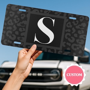 May include: A rectangular number plate featuring a dark grey and black leopard print pattern with a central black square containing a large white letter S. The plate has four mounting slots, one in each corner, and a smooth, flat finish.