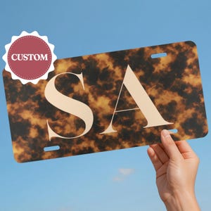May include: Custom license plate with a tortoiseshell pattern and the letters "SA" in a cream color. The word "CUSTOM" is printed on a red and white circular sticker.