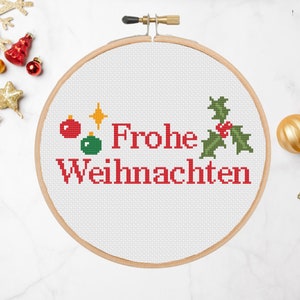 May include: A white embroidery hoop with a red and green cross-stitch design that says "Frohe Weihnachten" with a holly sprig and Christmas ornaments.