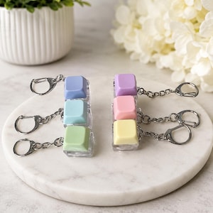 May include: Six individual keycap keychains arranged in two columns. Each keychain features a single square, pastel-coloured keycap attached to a silver-toned metal chain and lobster clasp. The colours include purple, pink, yellow, blue, teal, and green.