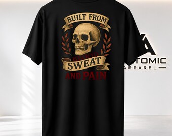 Puede incluir: Una camiseta negra de manga corta con un estampado gráfico de un cráneo humano centrado entre dos ramas con hojas. Encima y debajo del cráneo hay pancartas de color beige con el texto BUILT FROM, SWEAT y AND PAIN en letras rojo oscuro.