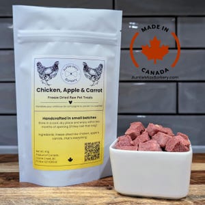Könnte beinhalten: Ein weißer Beutel mit gefriergetrockneten Rohfutter-Leckerlis mit der Aufschrift "Chicken, Apple & Carrot". Der Beutel zeigt Hühnerillustrationen und Text. Eine kleine weiße Schüssel enthält die Leckerlis. Hergestellt in Kanada.