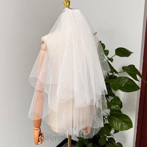 Könnte beinhalten: Ein weißer Tüll-Brautschleier mit einer einzigen Schicht und einem zarten Glitzern. Der Schleier wird auf einer Schaufensterpuppe präsentiert.