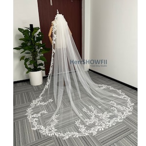 Könnte beinhalten: Ein weißer Braut Schleier mit einer zarten floralen Spitzenborte. Der Schleier ist lang und fließend, mit einem gewellten Rand. Das Spitzenmuster zeigt kleine Blätter und Blumen.