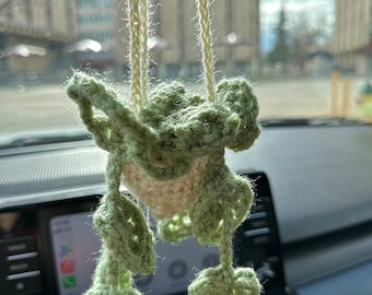 May include: A green crocheted hanging plant with white yarn strings. The plant is hanging from the rearview mirror of a car.