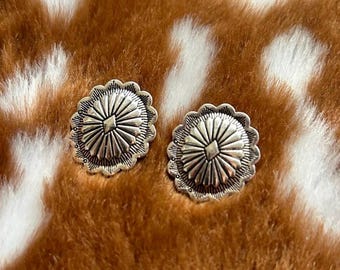 May include: A pair of silver-toned earrings with a circular design. The earrings have a raised, textured center with a starburst pattern.