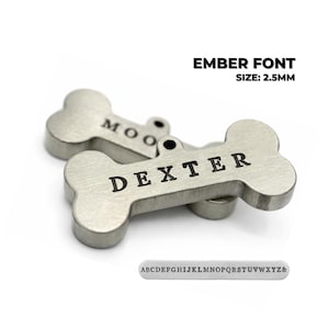Two silver-colored dog bone-shaped pet tags with engraved names. One tag reads "DEXTER" and the other reads "MOO". The text "EMBER FONT SIZE: 2.5MM" is above the tags. A small metal plate with the alphabet is below the tags. Ember font engraved dog tag personalized for pets showing custom dog tag engraving style