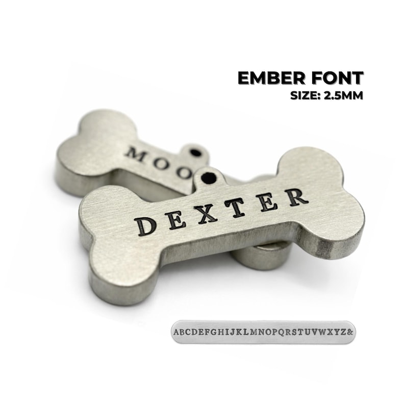 Two silver-colored dog bone-shaped pet tags with engraved names. One tag reads "DEXTER" and the other reads "MOO". The text "EMBER FONT SIZE: 2.5MM" is above the tags. A small metal plate with the alphabet is below the tags. Ember font engraved dog tag personalized for pets showing custom dog tag engraving style