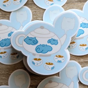 May include: A die-cut sticker featuring a white teapot with a light blue background. The teapot is decorated with blue cat-shaped clouds and is surrounded by small teacups containing steaming liquid and tiny blue cat figures.