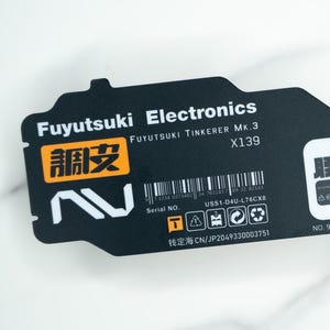 Peut inclure: Un composant électronique noir avec du texte blanc, dont "Fuyutsuki Electronics" et "Lithium polymer." Le composant a une forme unique avec un code-barres et divers symboles. Le texte "調皮" est visible dans un encadré orange.