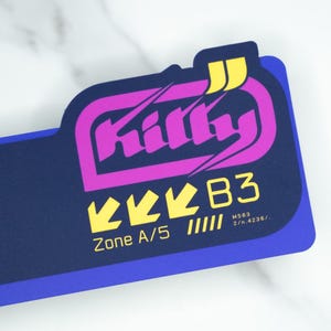 Puede incluir: Una pegatina rectangular con fondo azul oscuro y diseño morado y amarillo. La palabra "Kitty" está en una fuente estilizada. Debajo están las palabras "Zone A/5" y "B3".