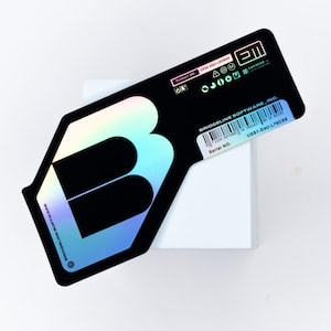 Könnte beinhalten: Ein schwarzer und schillernder Aufkleber mit einem stilisierten "B"-Design. Das "B" ist mit einem Farbverlauf aus Farben wie Blau, Grün und Lila gefüllt. Der Aufkleber hat Text und einen Barcode und befindet sich auf einer weißen Oberfläche.