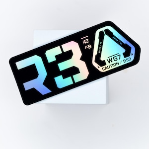 Może przedstawiać: Czarna prostokątna naklejka z iryzującym tekstem i grafiką. Naklejka zawiera liczby "R3" w dużej, stylizowanej czcionce, a także mniejszy tekst i trójkątny wzór. Tekst zawiera "WG7 CAUTION / B53".
