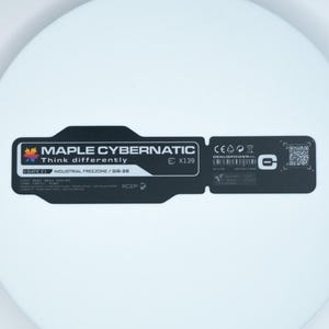 May include: A circular white disc featuring a central black rectangular label with rounded edges. The label displays the text MAPLE CYBERNATIC, Think differently, X139, GATE Z1, INDUSTRIAL FREEZONE, GIS-28, GENUSPOWER, and a QR code.