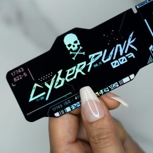 Puede incluir: Tarjeta negra con temática Cyberpunk con una calavera y huesos cruzados. La tarjeta presenta la palabra "CYBERPUNK" en una fuente futurista, junto con otros textos y símbolos. La tarjeta es sostenida por una persona.