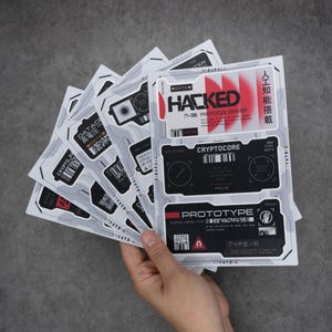 Puede incluir: Una pila de hojas de pegatinas rectangulares con gráficos futuristas de estilo industrial en blanco, negro y rojo. Los diseños incluyen texto técnico como HACKED, CRYPTOCORE y PROTOTYPE, junto con códigos de barras, códigos QR y caracteres japoneses.