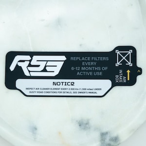 Puede incluir: Una pegatina rectangular en blanco y negro con bordes redondeados que presenta el texto RS3, REPLACE FILTERS EVERY 6-12 MONTHS OF ACTIVE USE, NOTICE, e INSPECT AIR CLEANER ELEMENT EVERY 2.500 Km (1.500 miles) UNDER DUSTY ROAD CONDITIONS.