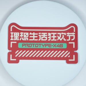 以下が含まれることがあります： 角が丸い長方形の赤いステッカー。白い中国語の文字、緑色の水平バーに書かれたPROTOTYPE-X48というテキスト、下部には白い斜めのストライプ模様があり、全体が白い枠線で囲まれています。
